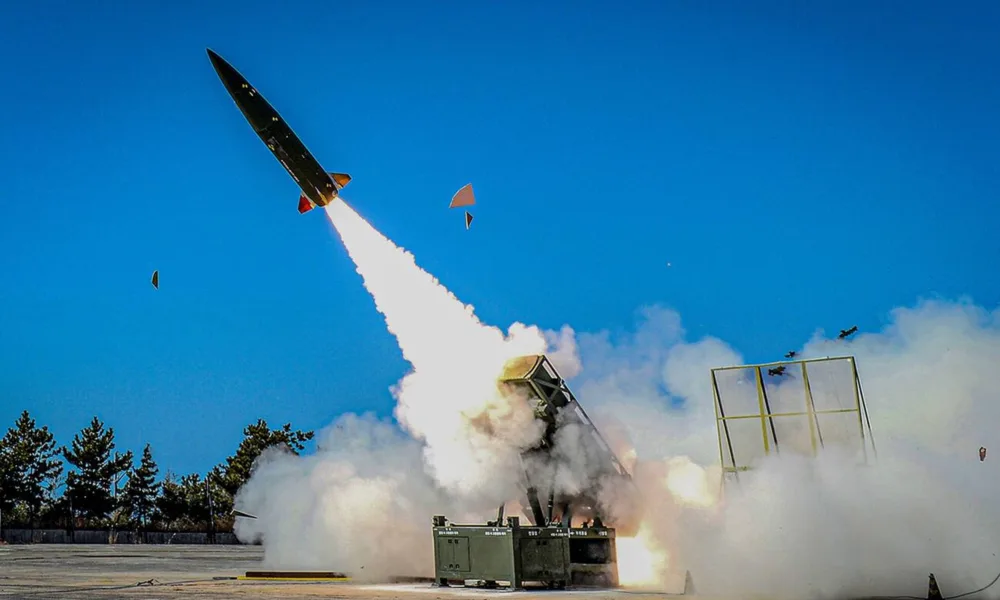 A modern missile being launched at high speed from a ground launcher during a test, with a blue sky and clouds of smoke surrounding the site.