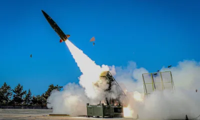 A modern missile being launched at high speed from a ground launcher during a test, with a blue sky and clouds of smoke surrounding the site.