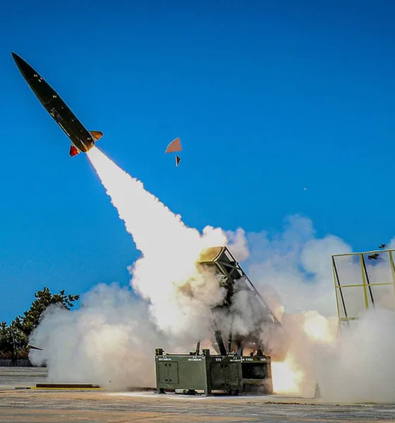 A modern missile being launched at high speed from a ground launcher during a test, with a blue sky and clouds of smoke surrounding the site.